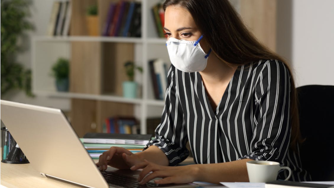 A woman with a facemask on, typing on her laptop