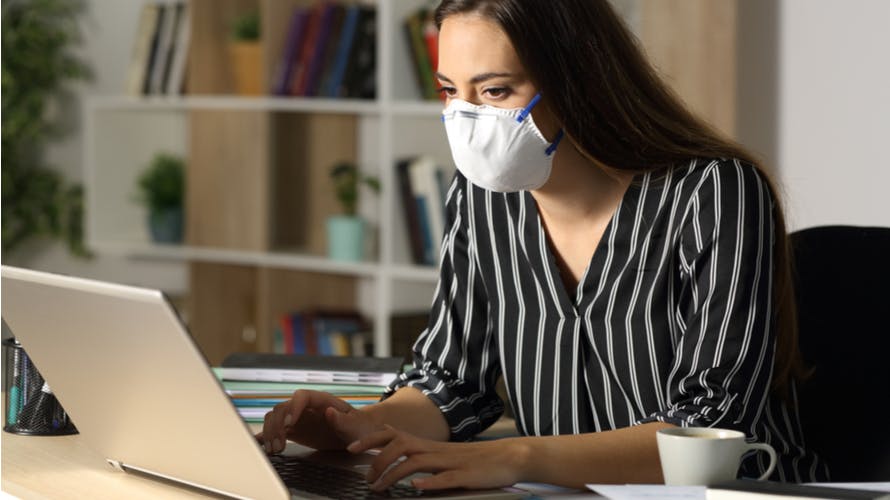A woman with a facemask on, typing on her laptop