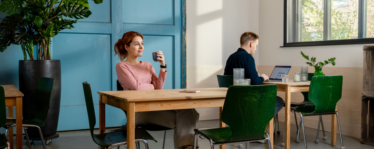 Ondernemer aan het werk achter de laptop op een coworking plek. Andere ondernemer even niets aan het doen met een glas drinken in haar hand