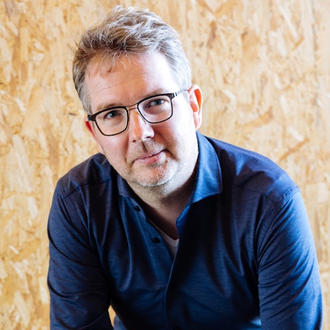 Joost kijkt lachend in camera voor houten achtergrond