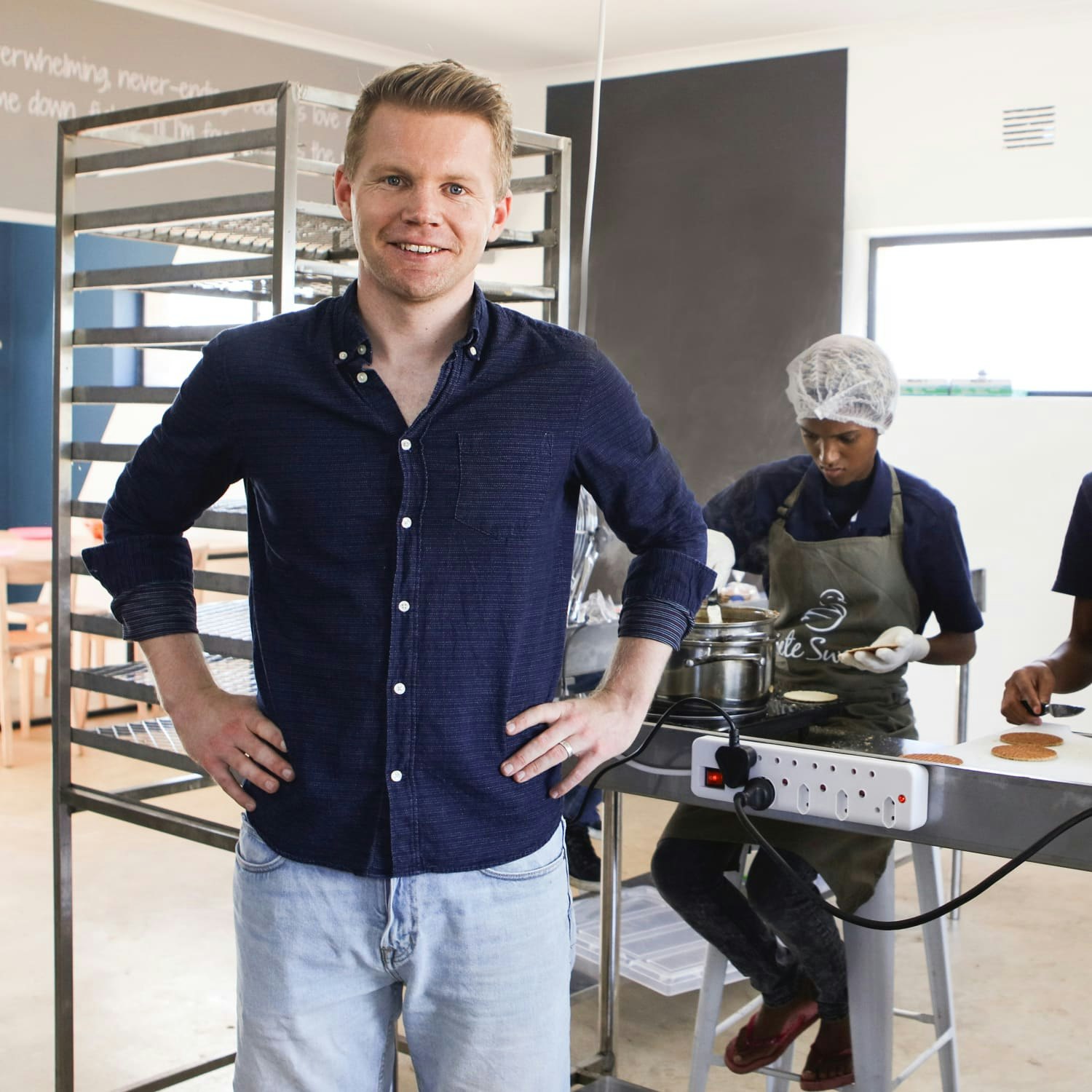 Derk kijkt lachend in de camera terwijl collega's stroopwafels maken