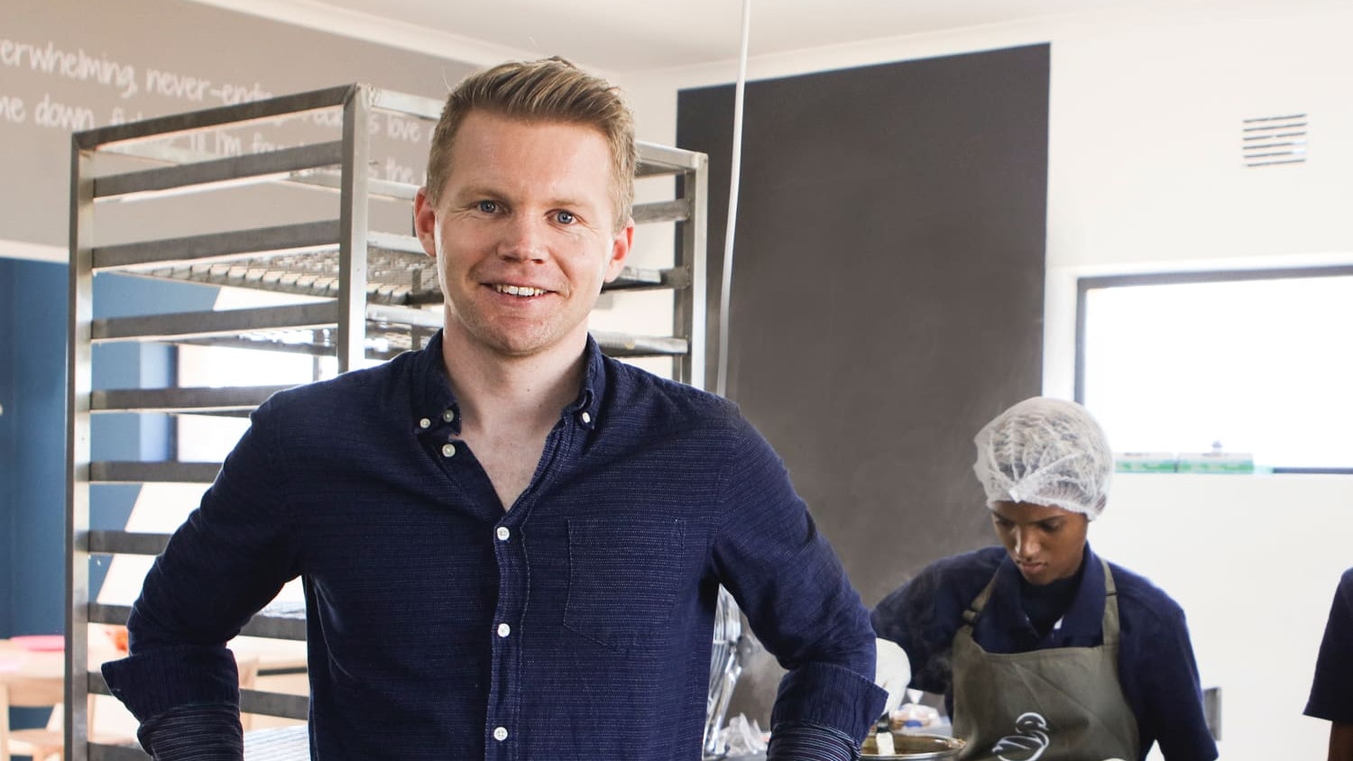 Derk kijkt lachend in de camera terwijl collega's stroopwafels maken