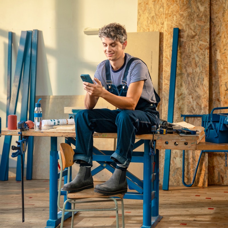 Timmerman op zijn telefoon in zijn werkplaats met blauw overall aan