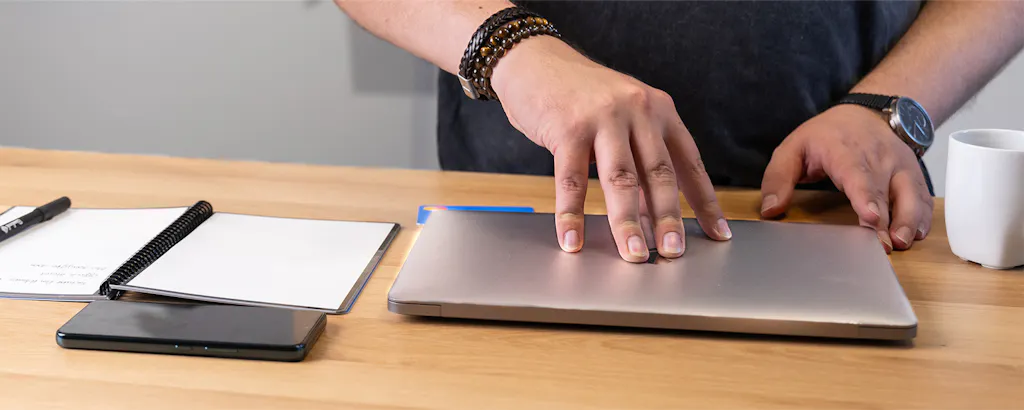 Iemand die zijn laptop open wil gaan klappen met koffie, notitieblok en telefoon op tafel
