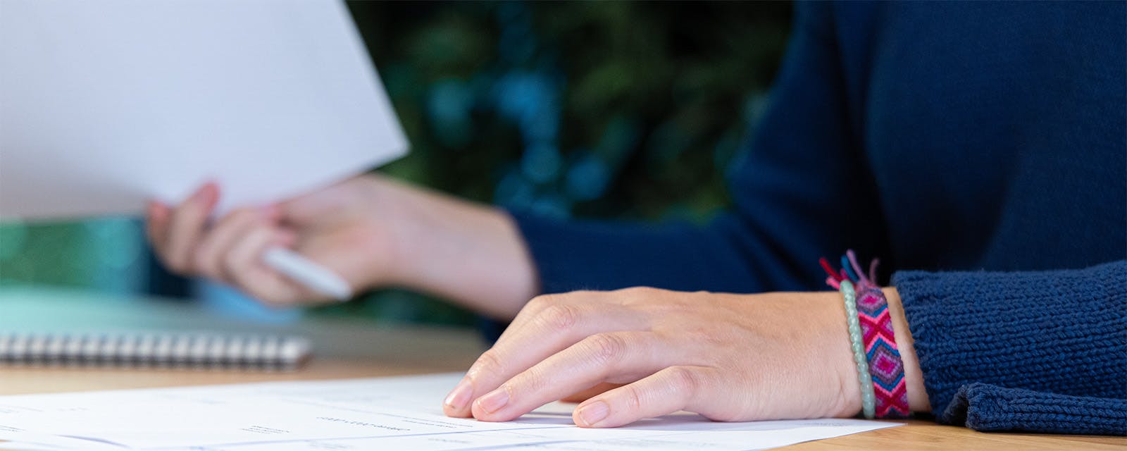 Ingezoomde foto van linker hand op stapeltje papieren en rechterhand houdt een papier en pen vast