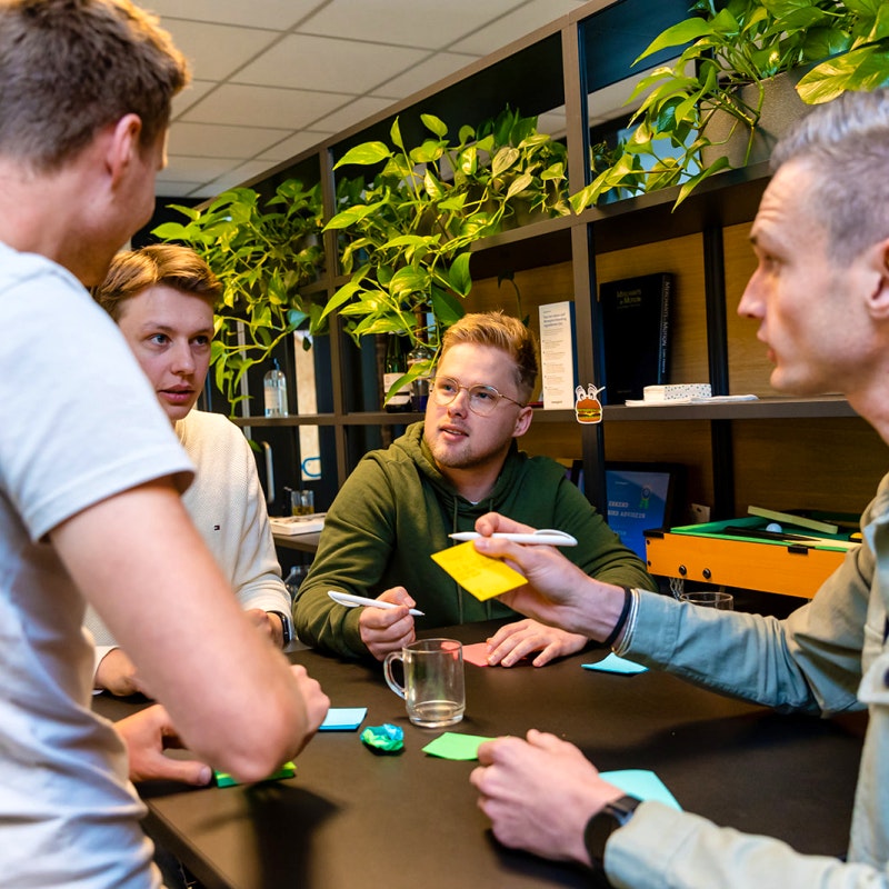 Tim, Jeroen, Sven en Gijs aan het brainstormen in de kantine tijdens een kennissessie met kastwand en planten op de achtergrond