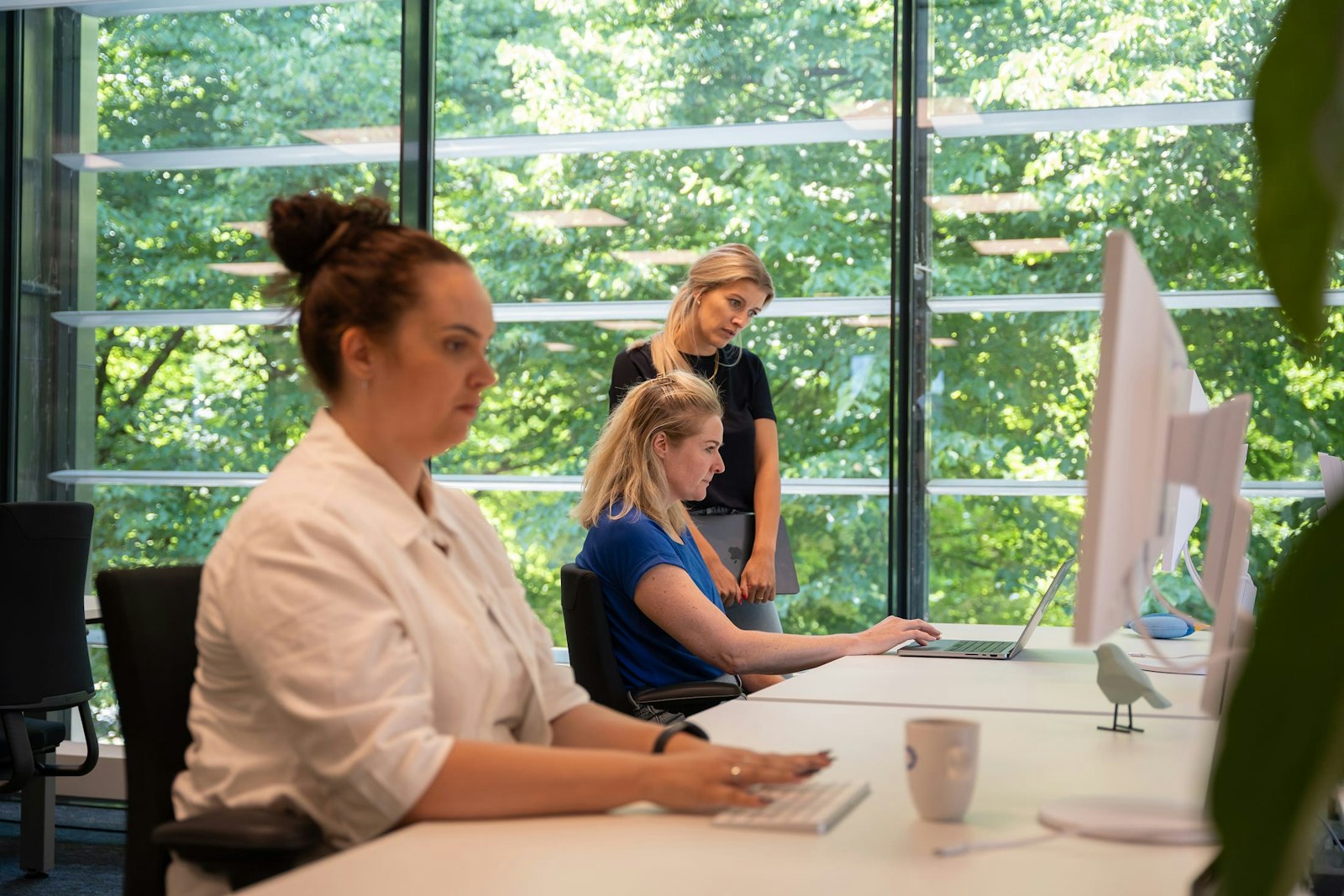 Suzan kijkt bij Alieke op haar scherm, Nathalie is op de voorgrond aan het werk