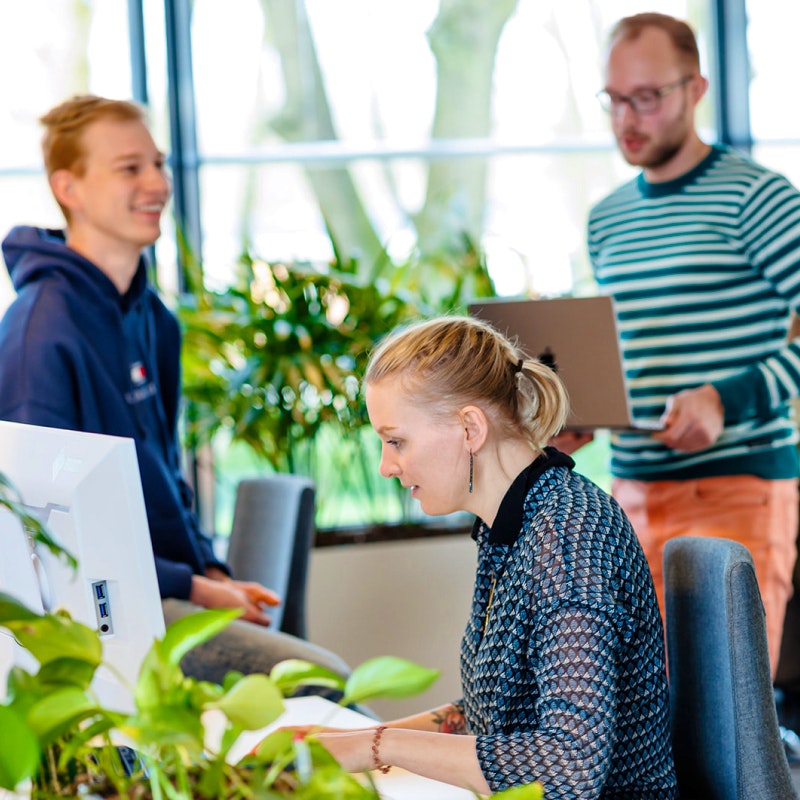 Op kantoor waar Loes achter haar laptop zit, Jeroen met zijn laptop loopt en Jesper een gesprek voert met een collega buiten beeld