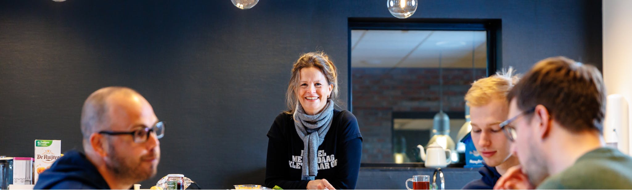 Margo kijkt in camera vanaf de bartafel in de kantine terwijl andere collega's op de voorgrond aan het lunchen zijn
