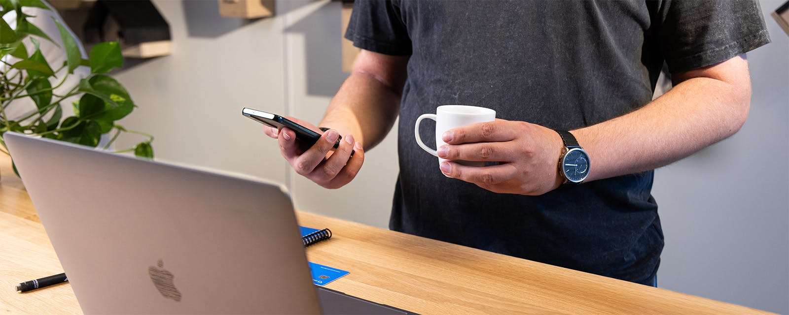 Uitsnede van persoon met laptop en koffie in de hand terwijl hij achter een hoge tafel staat met zijn laptop voor zich