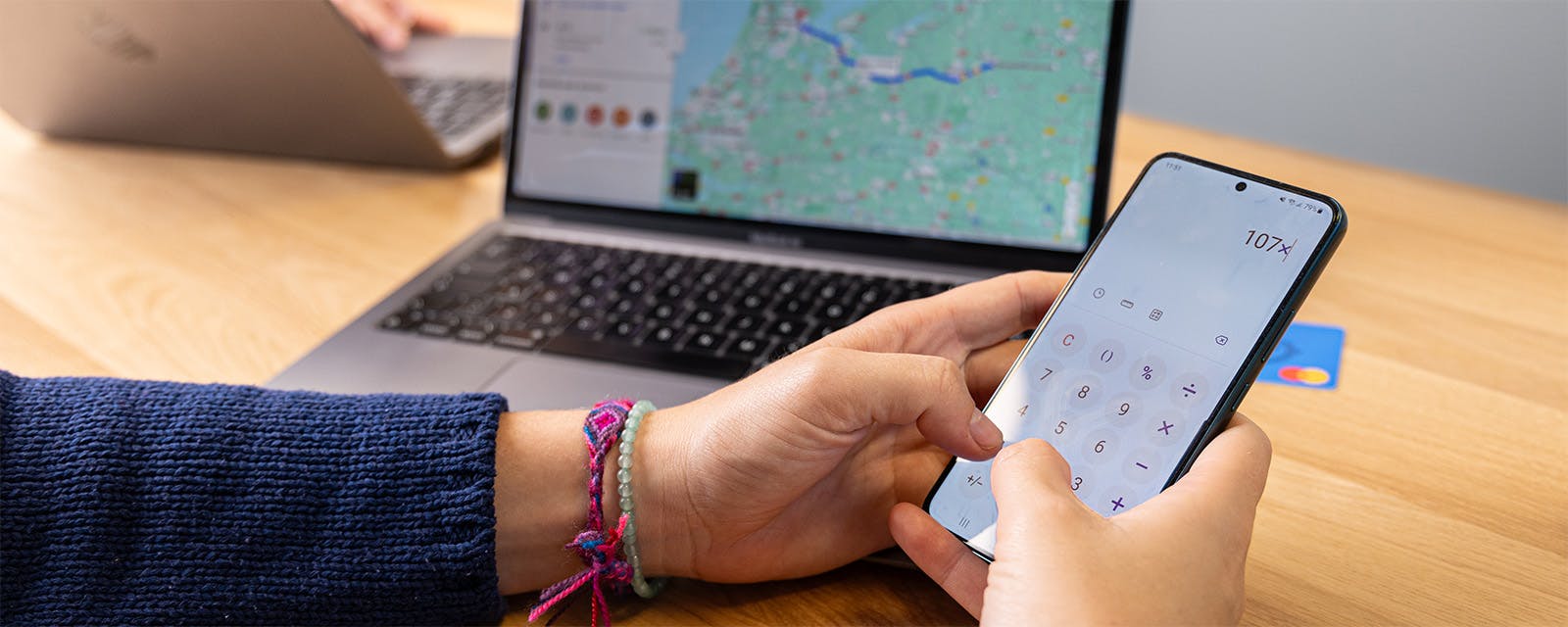 Close-up van handen die op een mobiel een berekening maken van het aantal gereden kilometers met op de achtergrond een routeplanner