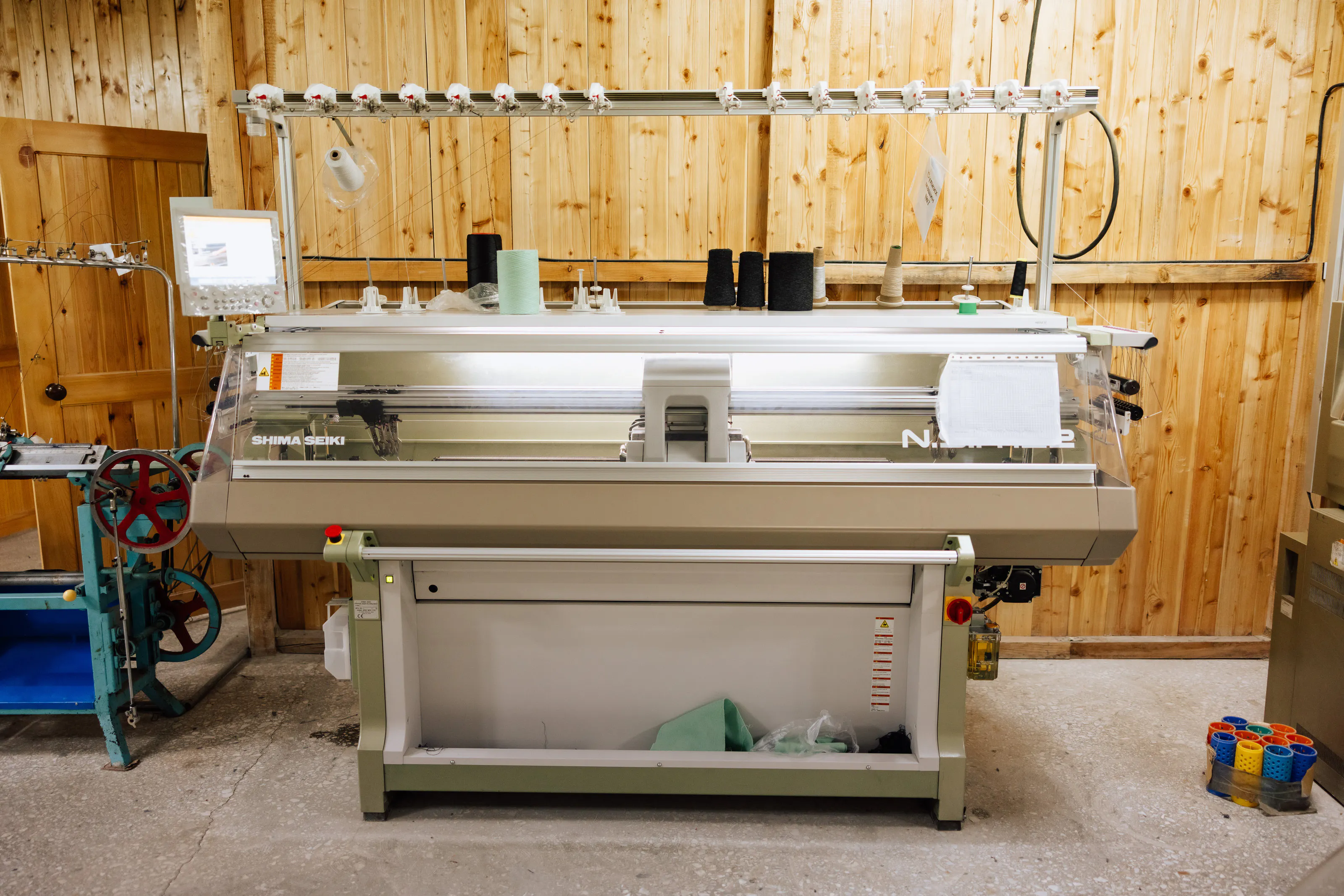 A flat knitting machine with yarn cones above it.