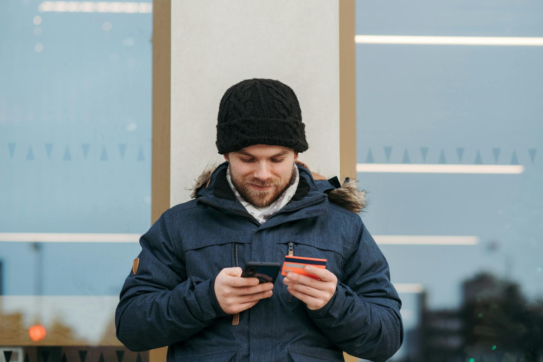 Homem com capuz e jaqueta segura o smartphone numa mão e o cartão de crédito na outra