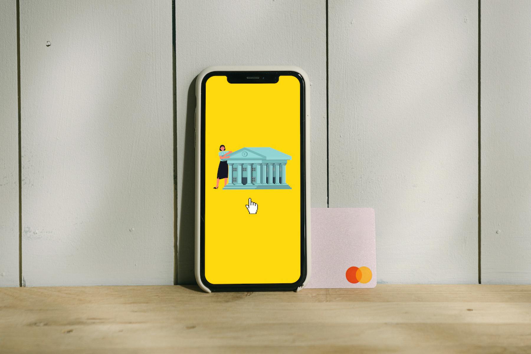 Smartphone mostra uma ilustração de banco digital ao lado de um cartão de crédito sobre uma mesa de madeira e parede branca ao fundo