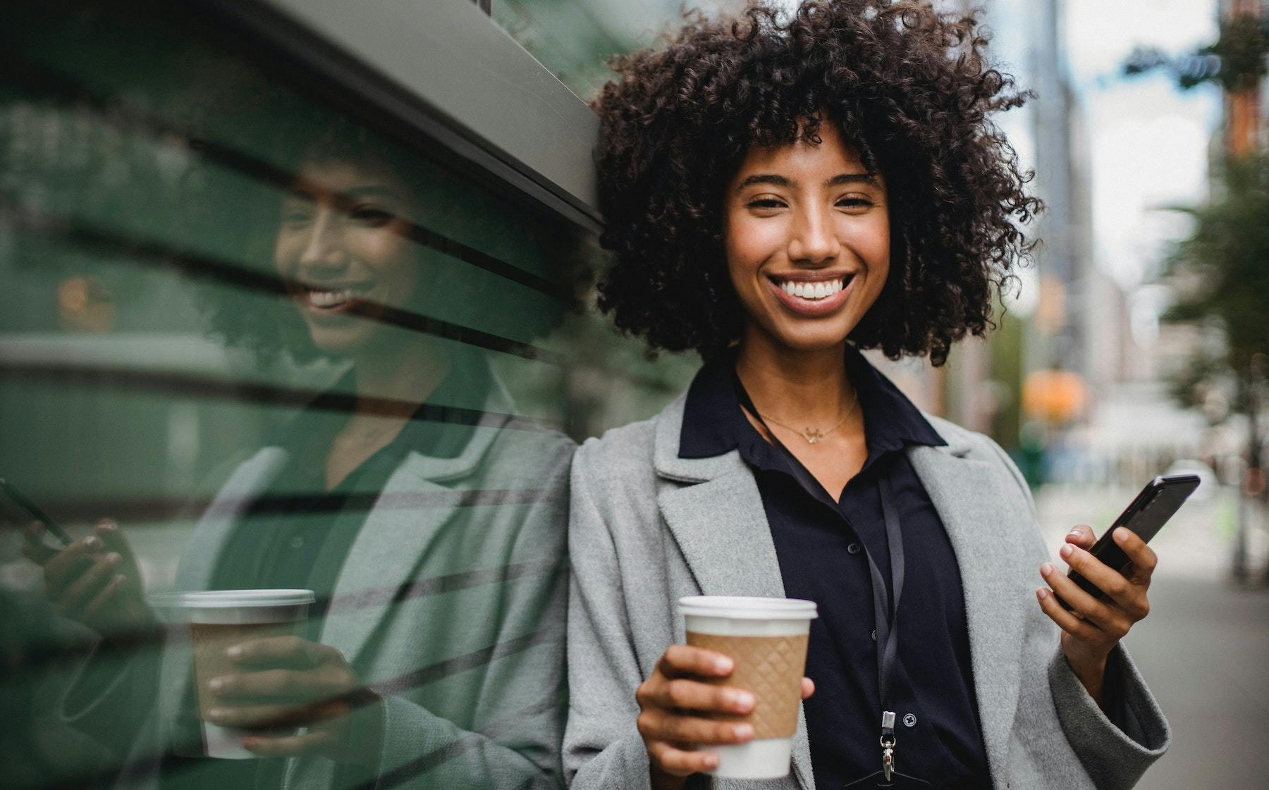 Mulher negra sorri enquanto segura o celular e um copo de café