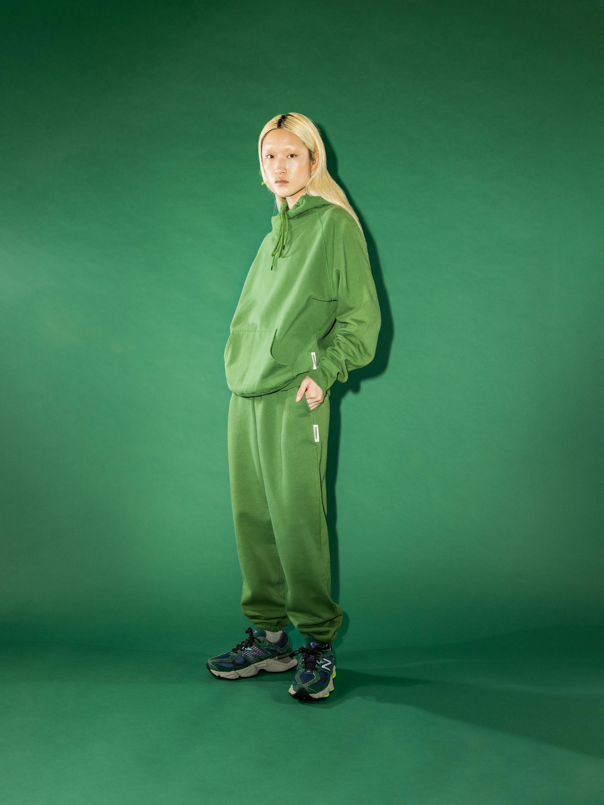 Weibliches Model trägt grünen Hoodie und Joggingshose von Highsnobiety vor grünem Hintergrund.