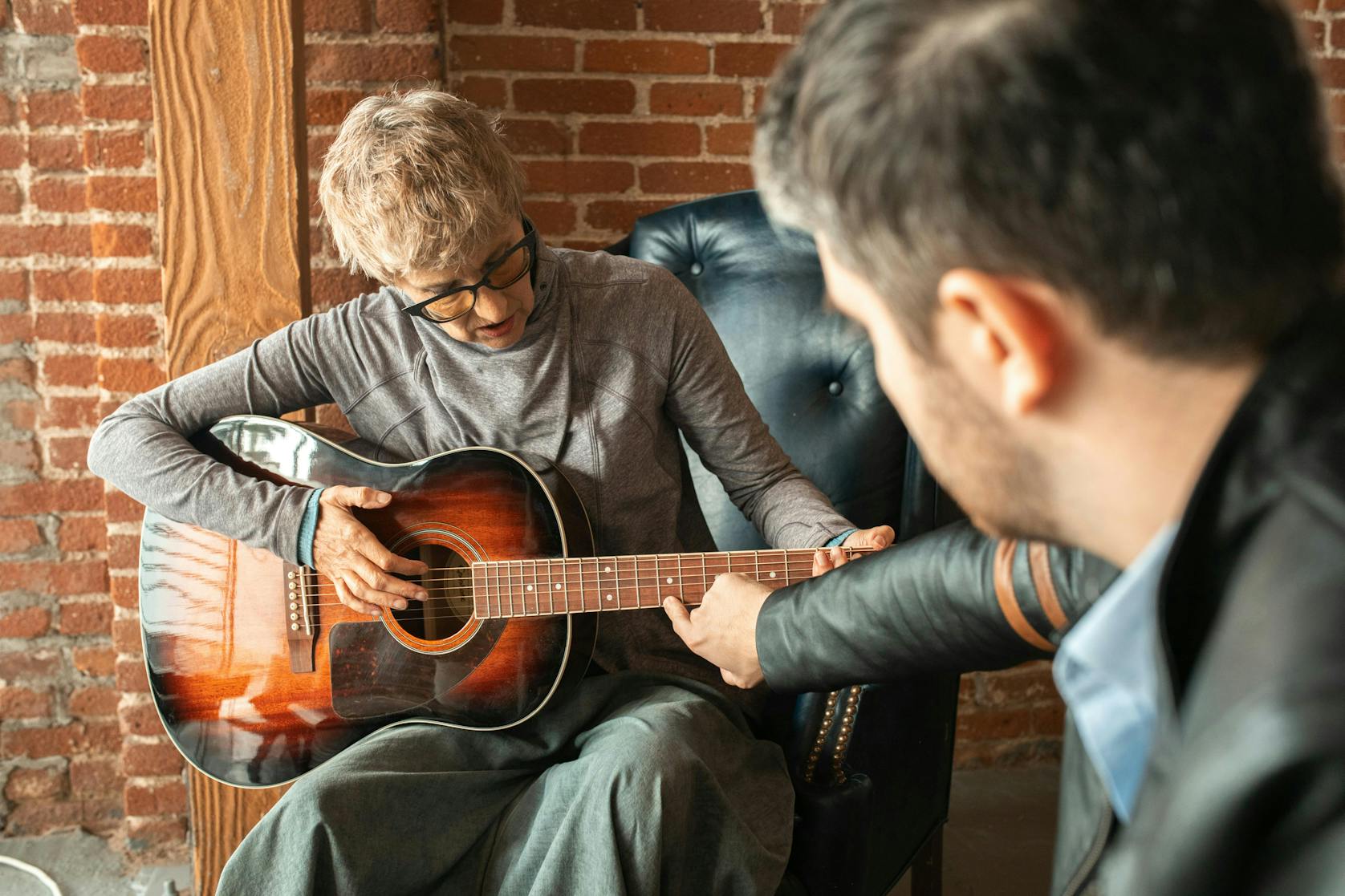 An adult woman playing the guitar while her instructor gently guides her finger placement on the fretboard during a lesson