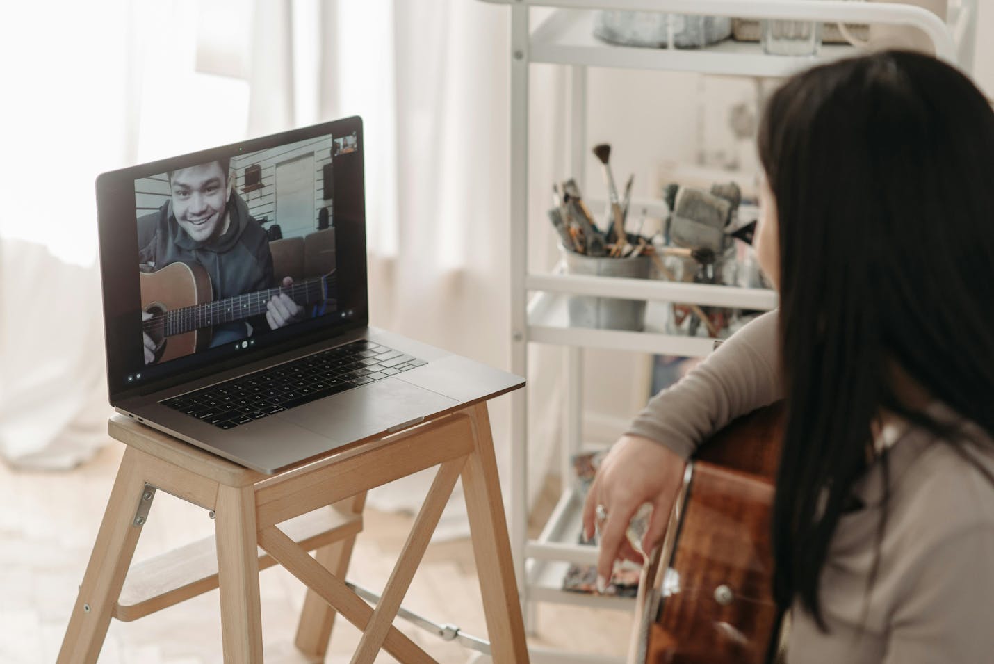 A student has her guitar on her lap and is sitting in front of her computer while she is talking to her guitar teacher who is also holding his guitar. They are having online guitar lessons.