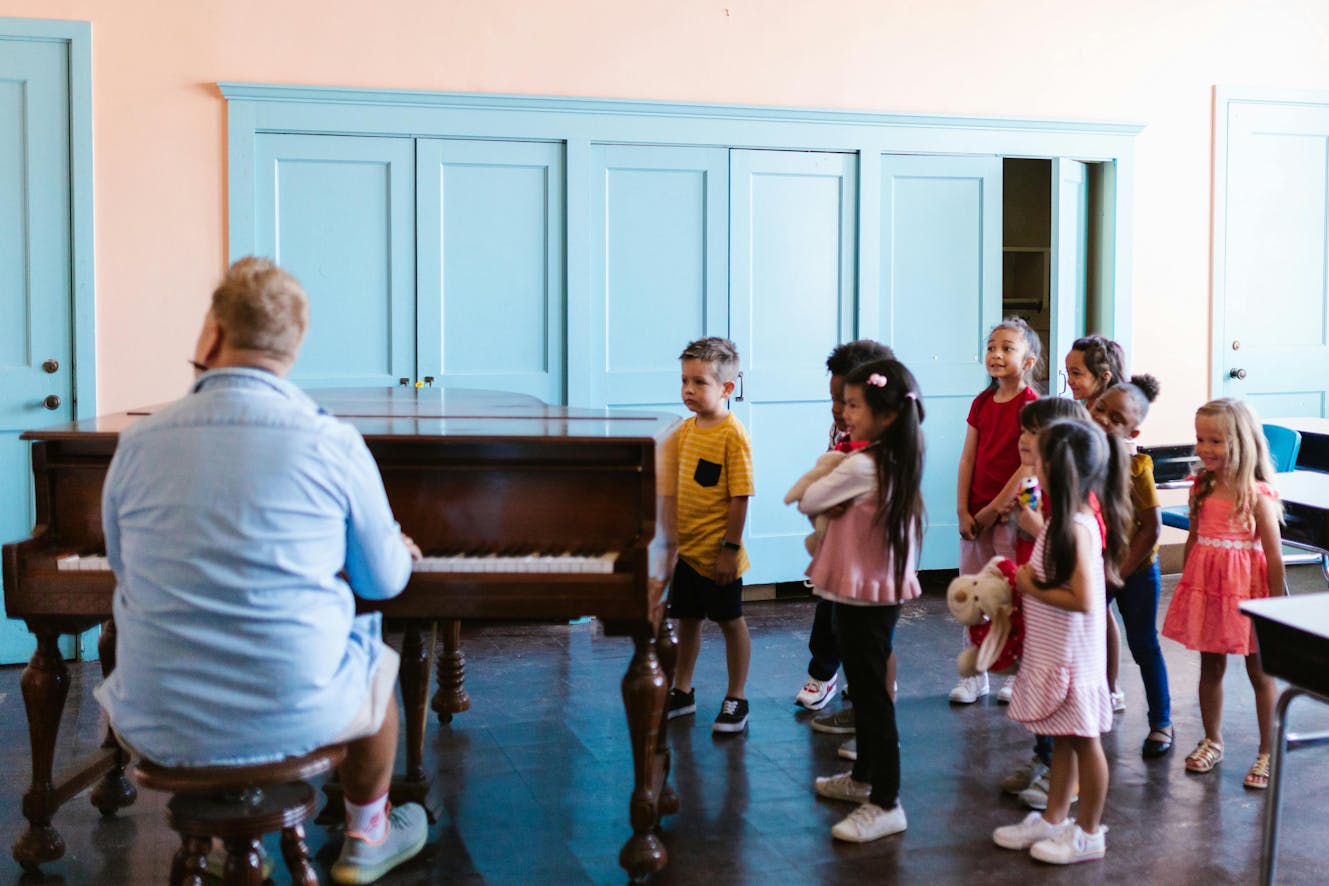 Nine kids face the piano and sing along while the music teacher plays and teaches kids in school.