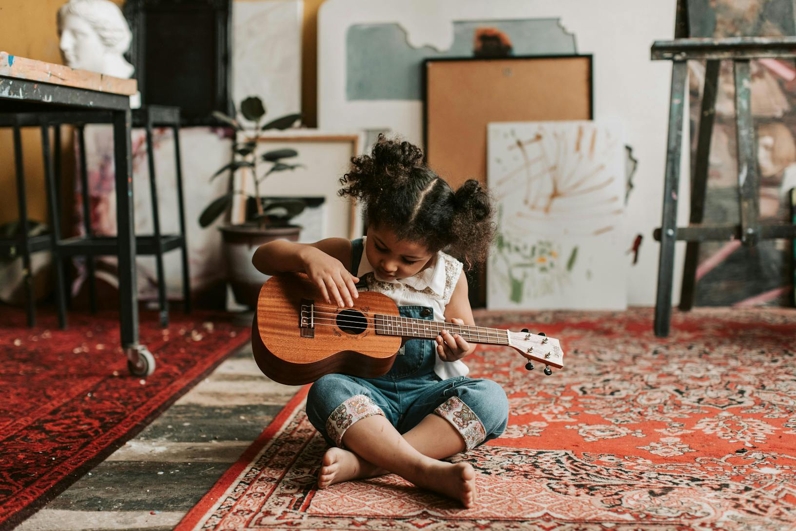 A preschooler sitting on the floor holding a ukulele, learning to play.