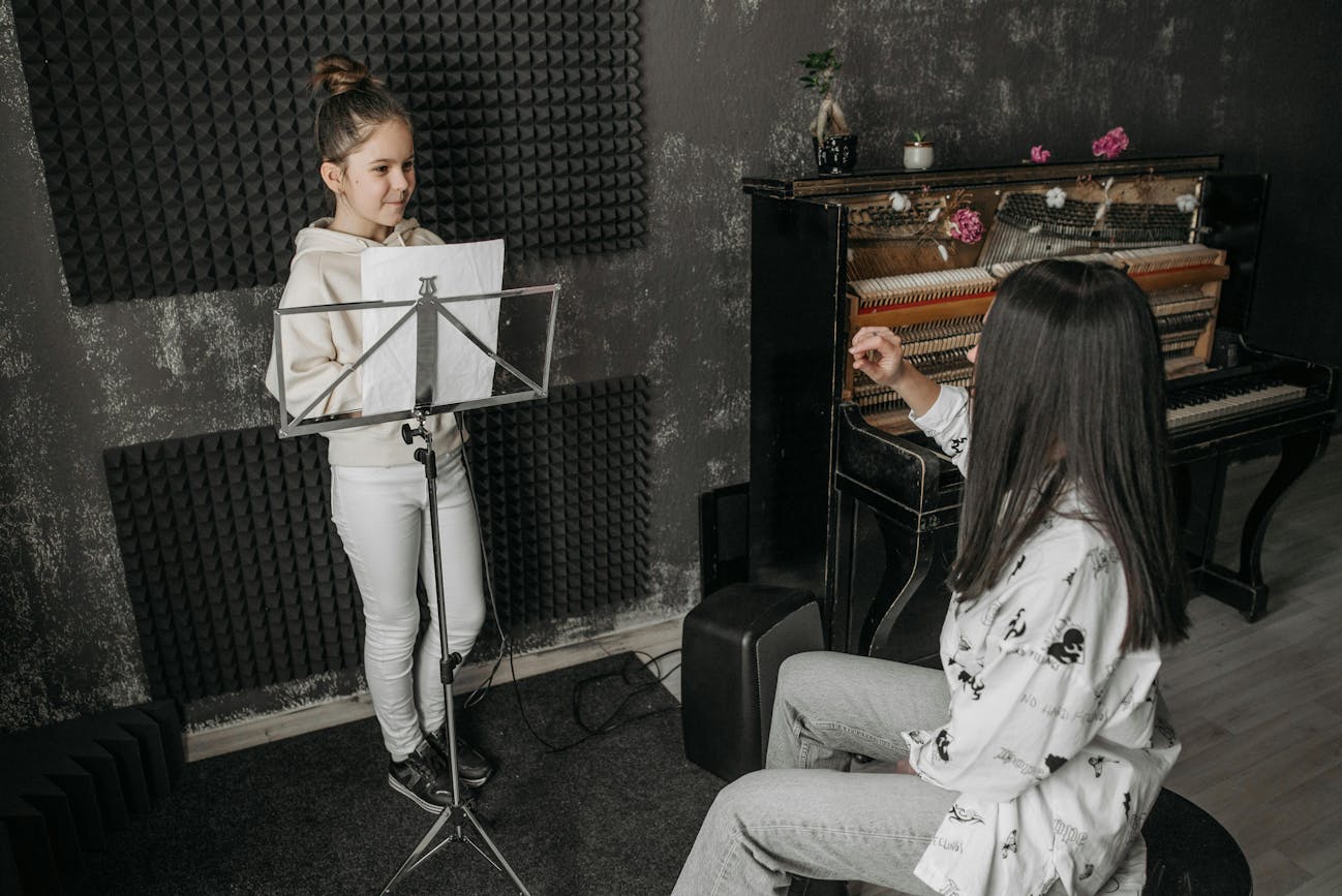 A young girl in a white hoodie and pants stands at a music stand, reading sheet music, while a woman with long dark hair in a patterned white shirt sits nearby, is teaching her. They are in a music room with soundproofing panels on the walls and an upright piano decorated with flowers in the background