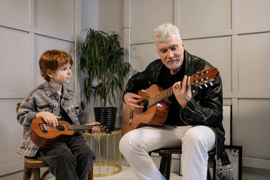 A young boy playing a ukulele sits beside an older man playing a classical guitar. The boy wears a denim jacket and looks attentively at the man, who is dressed in a black leather jacket and white pants. They are seated in a bright room with a plant and framed artwork in the background