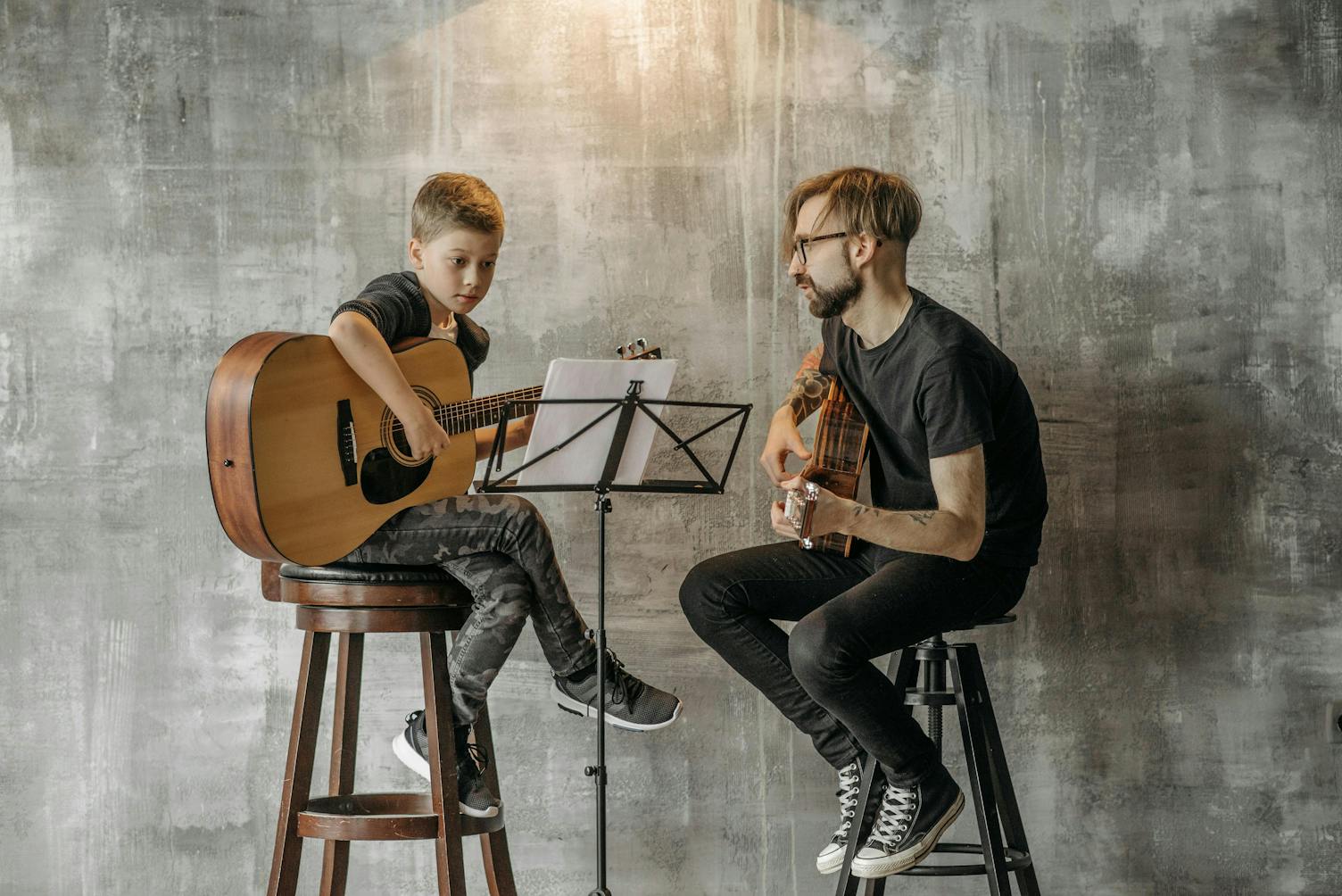 A young boy and an adult man are seated on high stools, both playing acoustic guitars. The boy is focused on reading sheet music placed on a stand between them. The man teaching the boy watches attentively.