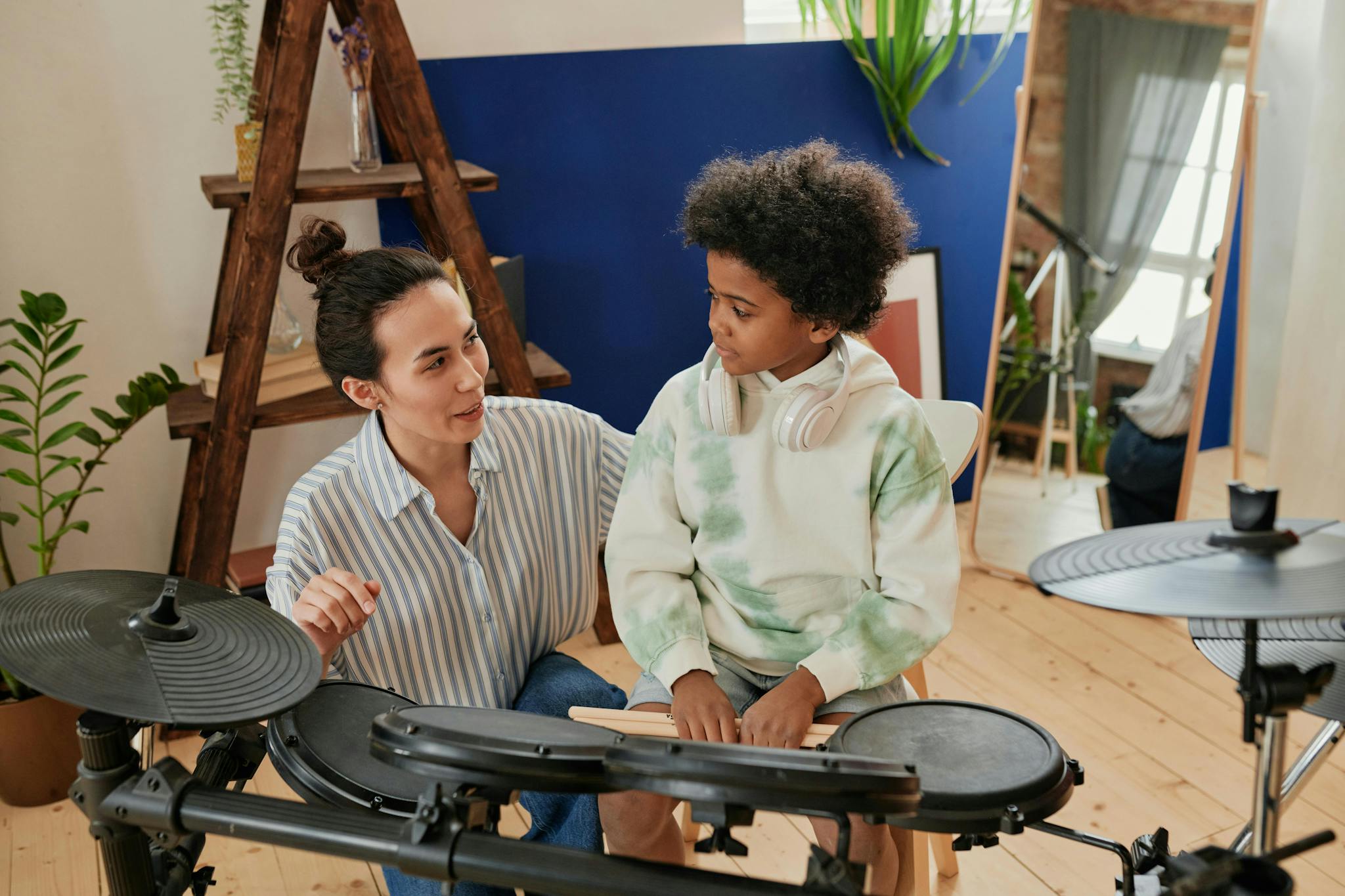 A music instructor teaching a young student how to play an electronic drum set in a cozy studio. The instructor is gesturing and smiling, while the student listens attentively, holding drumsticks and wearing headphones around their neck