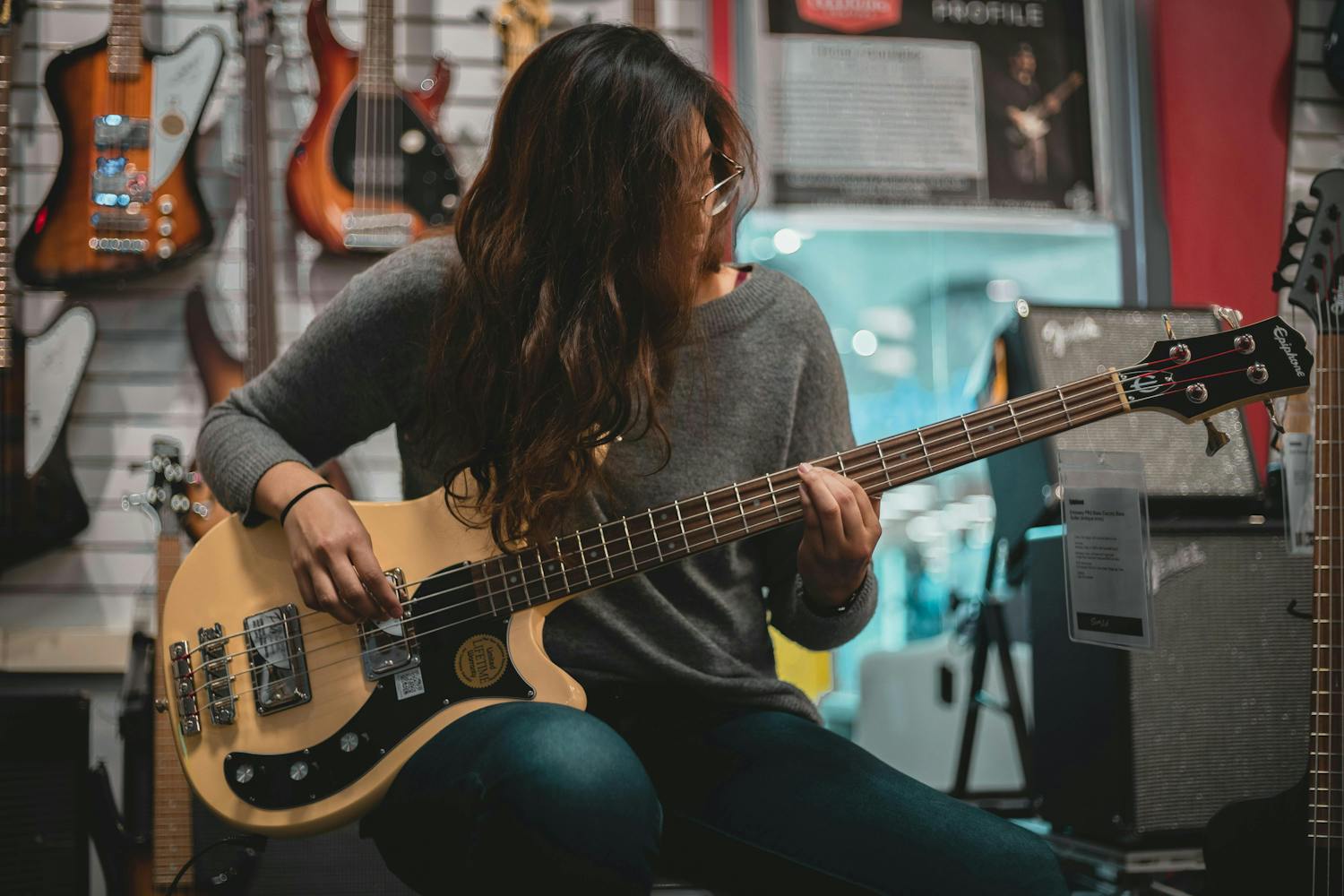 A woman playing a bass guitar, surrounded by guitars hanging on the wall and amplifiers on display. She is focused on learning to play.
