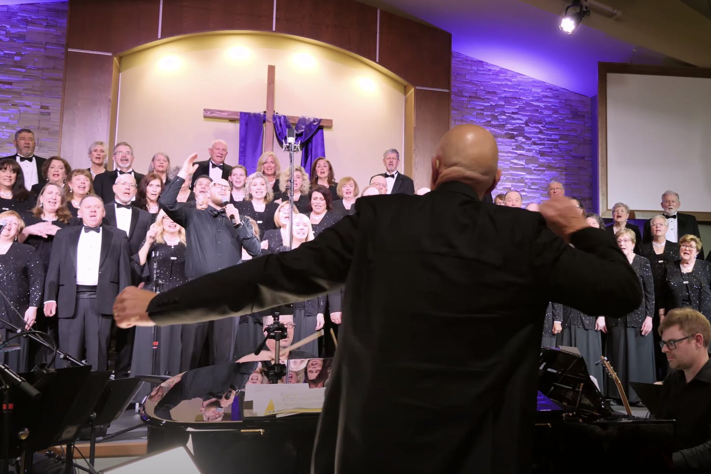 Allan Skoog directing the mosaic choir in a church with a cross behind them