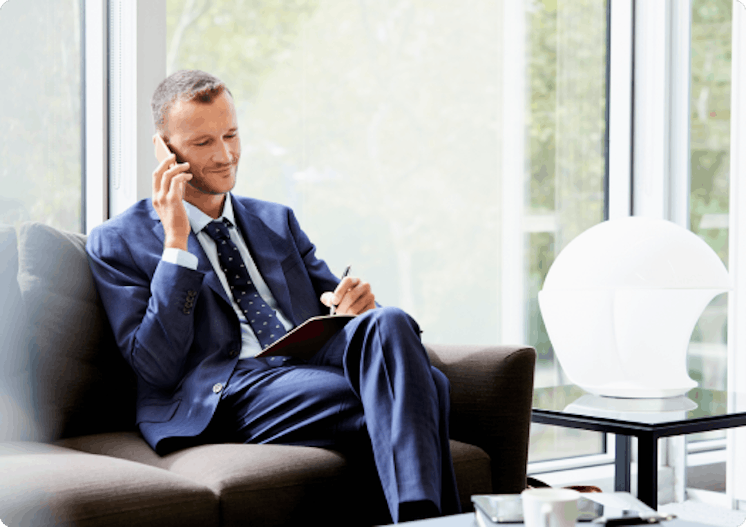 A man in work attire speaks on the phone whilst sitting on a sofa
