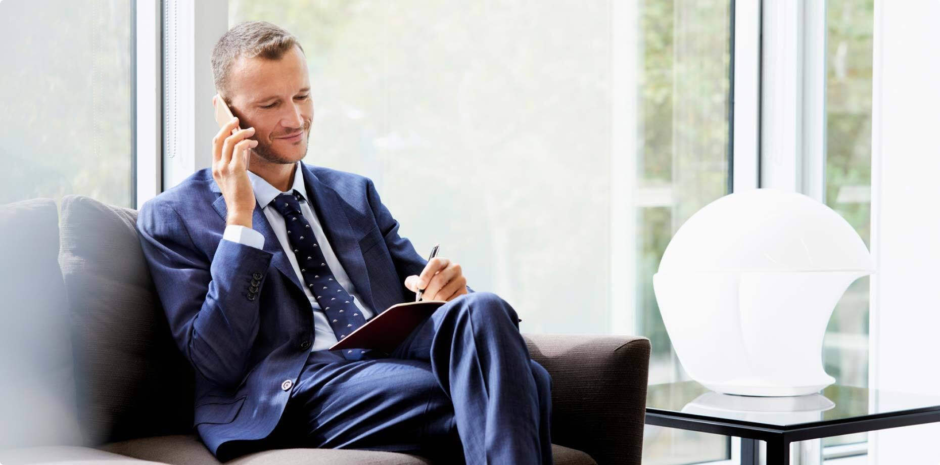 A man in work attire, sitting on a sofa and talking on the phone