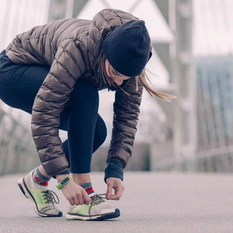 A lady in a beanie, puffer jacket and running leggings, is lacing up her trainers before running or walking to work.