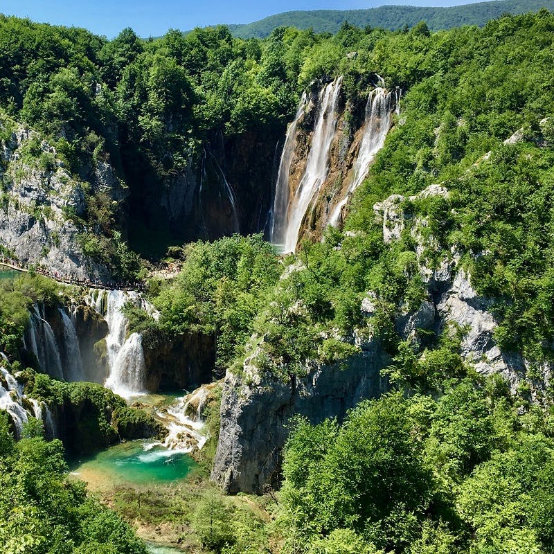 A large waterfall of 2 steps ending in a turquoise body of water. The waterfall is surrounded by lush green vegetation. 