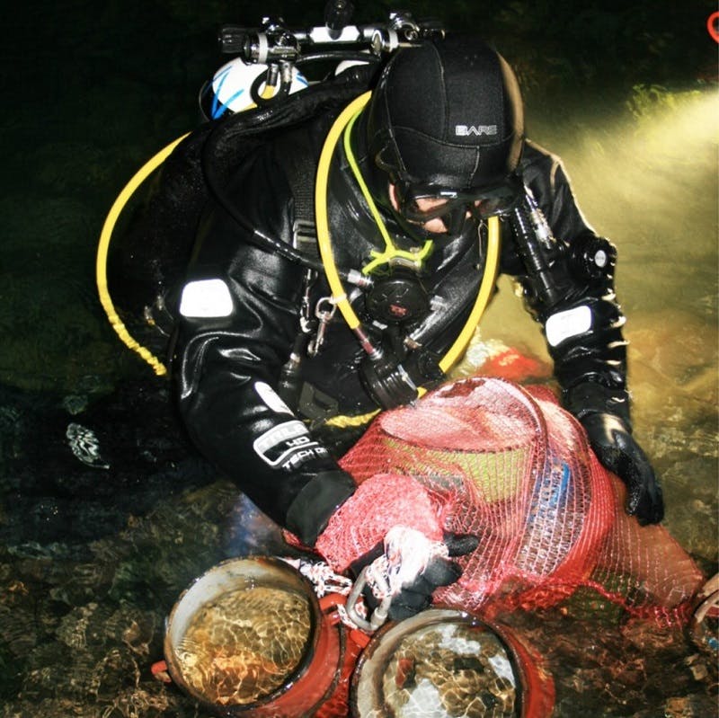 A cave diver partially out of water, with full equipment, putting solid waste into a bag inside a cave. Credit: PROTEUS project team.