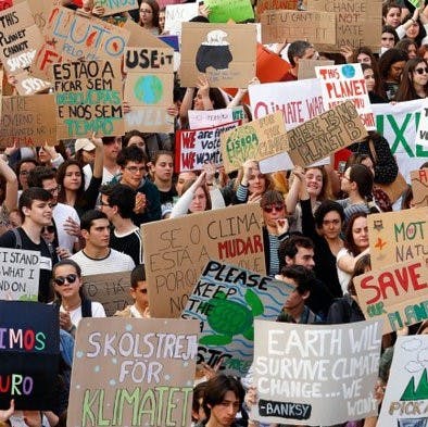 A group of young people at a climate strike, one holding a sign that reads "Less is More. It's Eco-Logical"