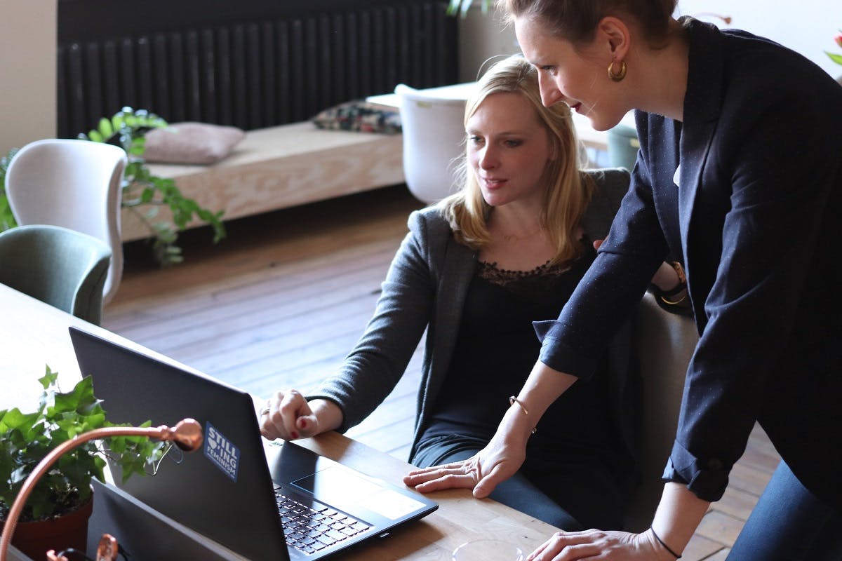 Two women looking at a computer screen in a bright open spaced office. Social interaction and brainstorming is one of the cons of of working from home. 