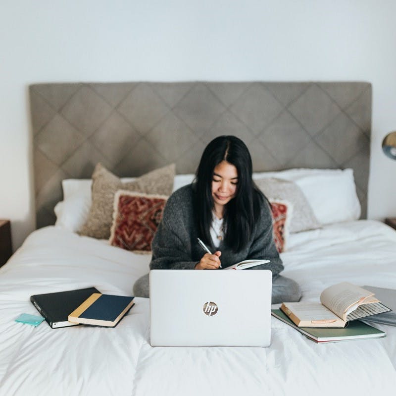 A woman enjoying her work from home perk with her laptop and books on her bed.