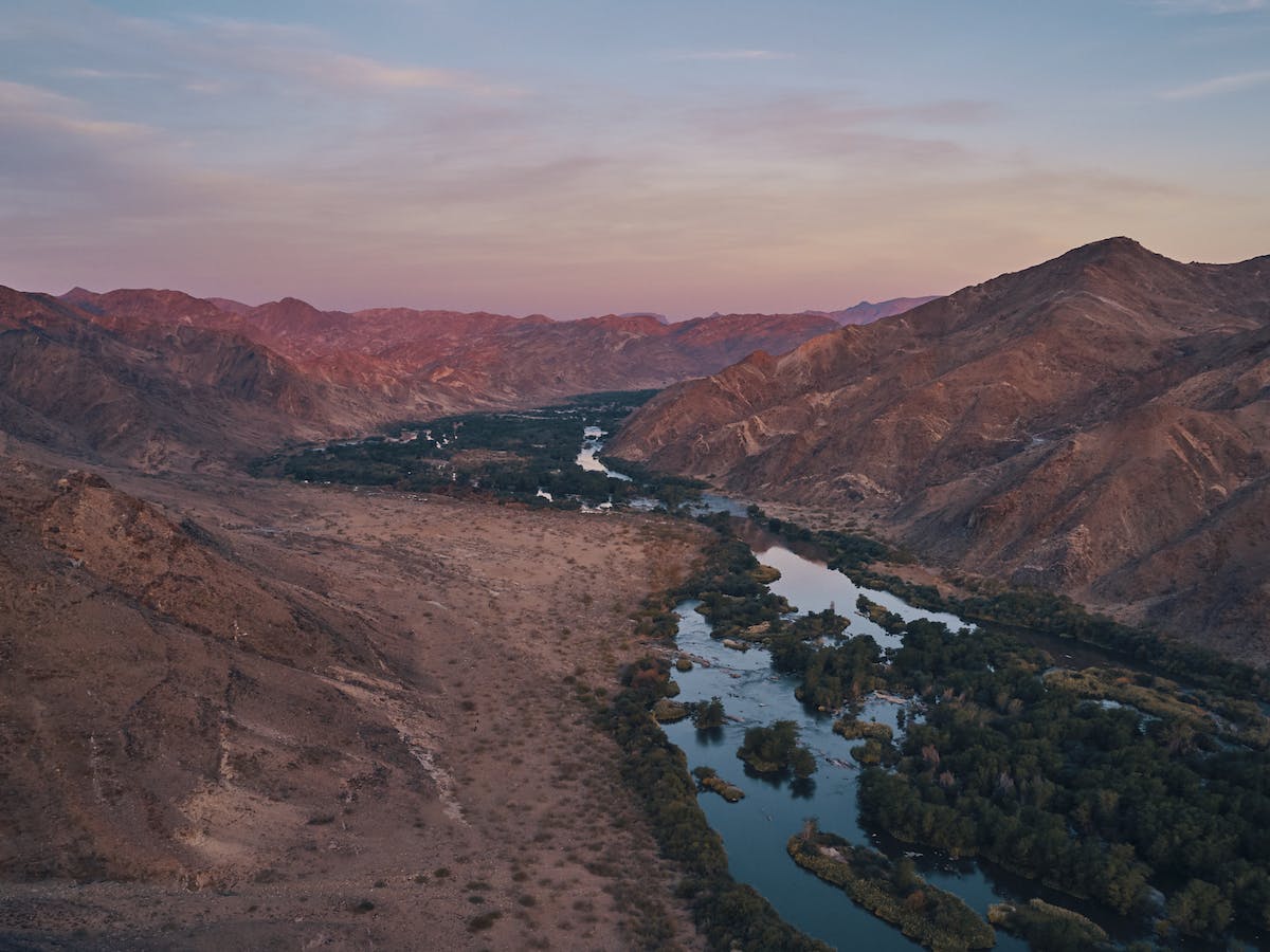 A lush river runs through mountains in the centre of the image, with pink sunset hues filling the sky