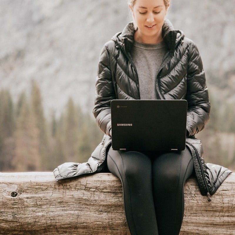A woman sitting on a log working on her laptop with nature in the background.