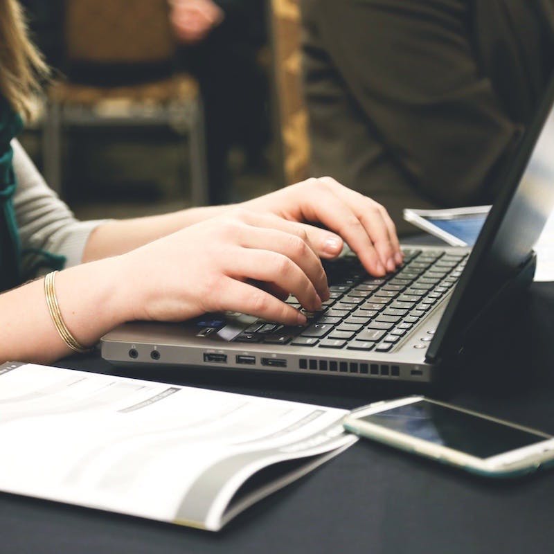 A woman types onto a laptop with her phone beside her. 