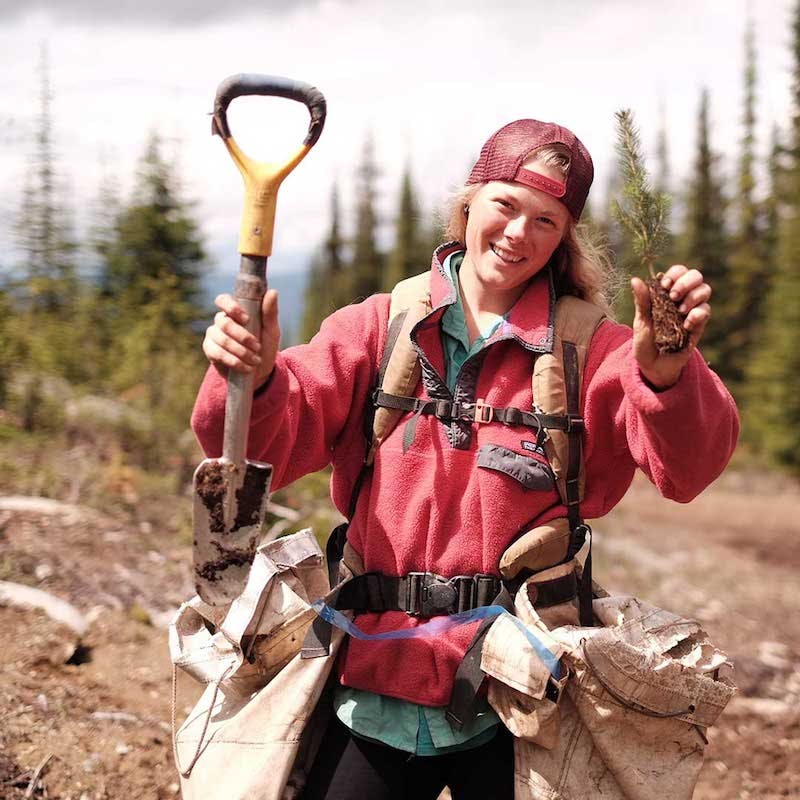 A tree planter in the field with a shovel and tree sapling in each hand. Find out her story at Patagonia's Worn Wear story share.