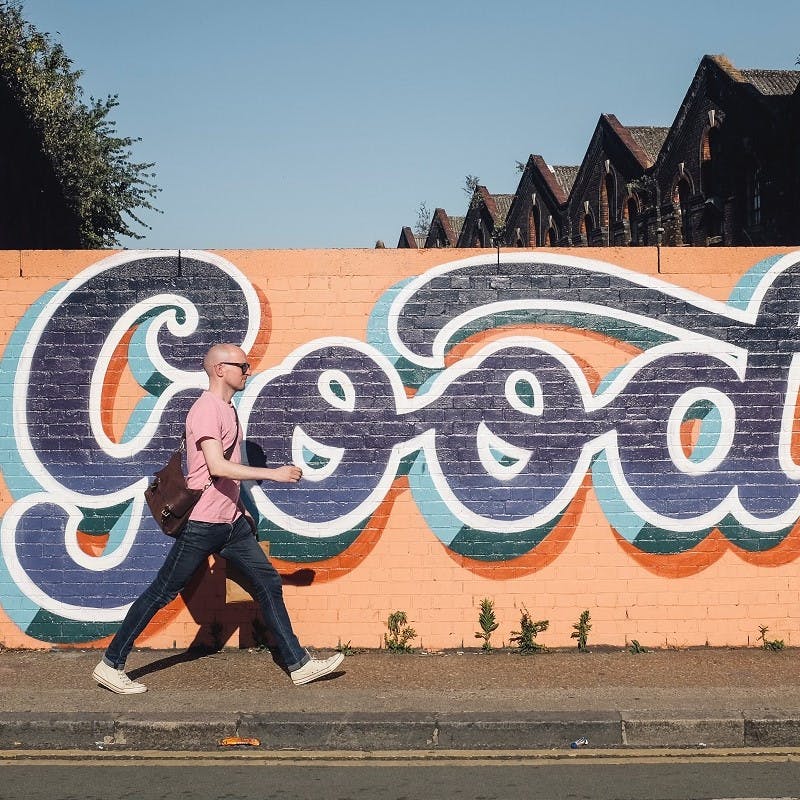 A man walking to work, past a wall with bright blue and orange graffiti reading "good".