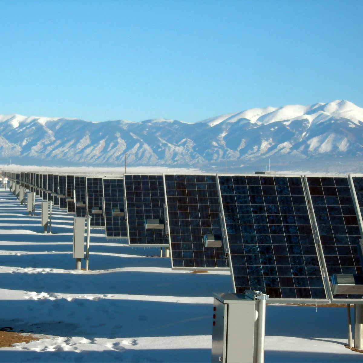 A row of solar panels under blue skies  in front of snow capped mountains