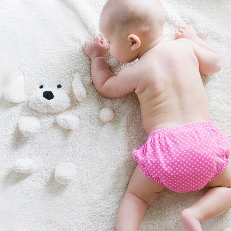 A baby dressed in a pink reusable cotton nappy next to its teddy bear. 