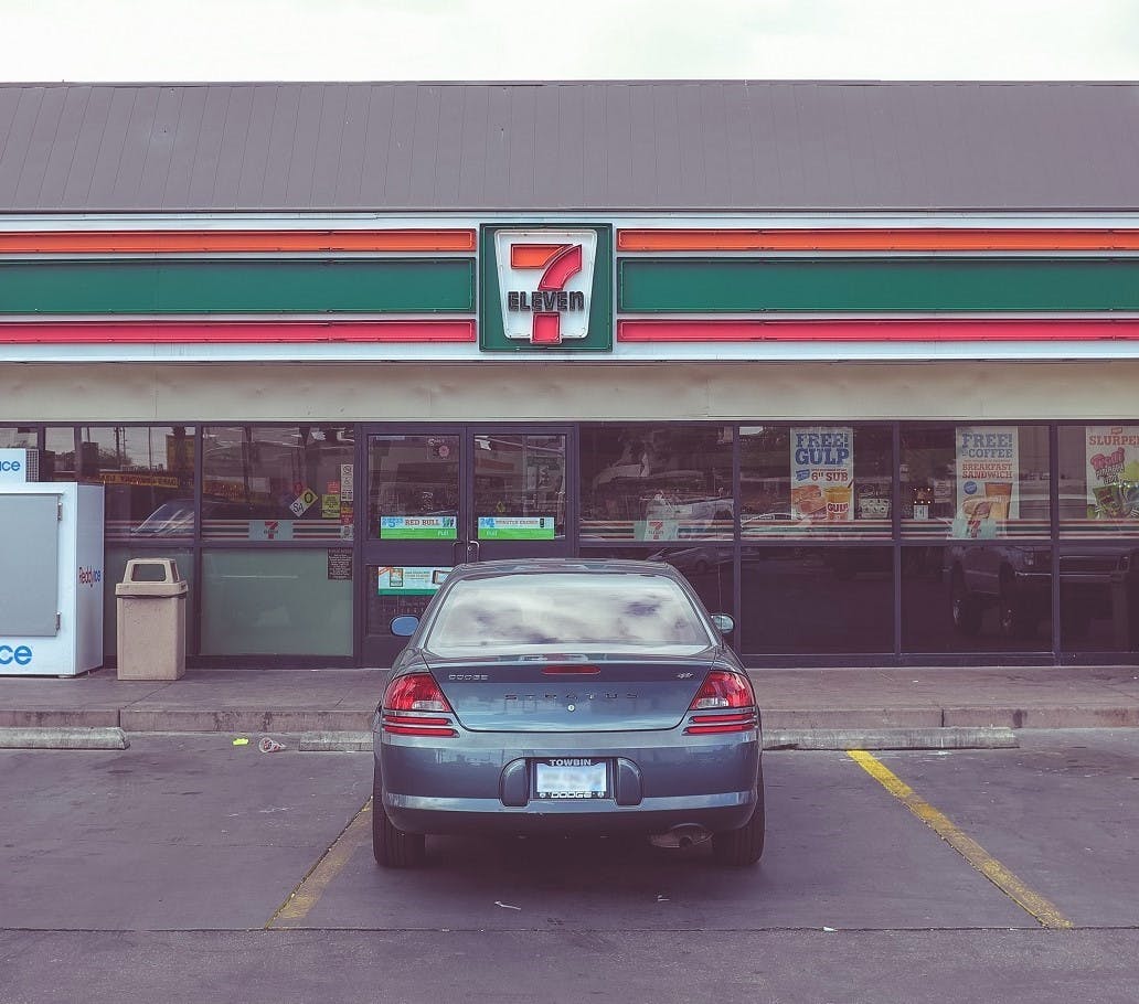 A silver car parked outside of a 7 Eleven convenience store. Walking, running or cycling to the convenience store can low your carbon footprint. 