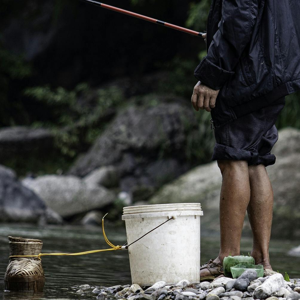 A fisherman by a stream with a rod and bucket.