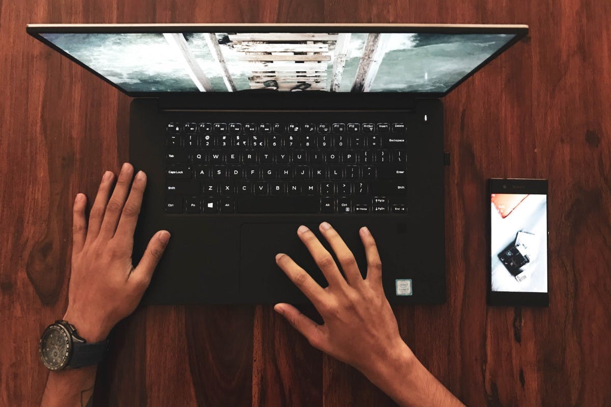 Bird's eye view of someone typing on their computer on a wooden table with a smartphone lying on the side.