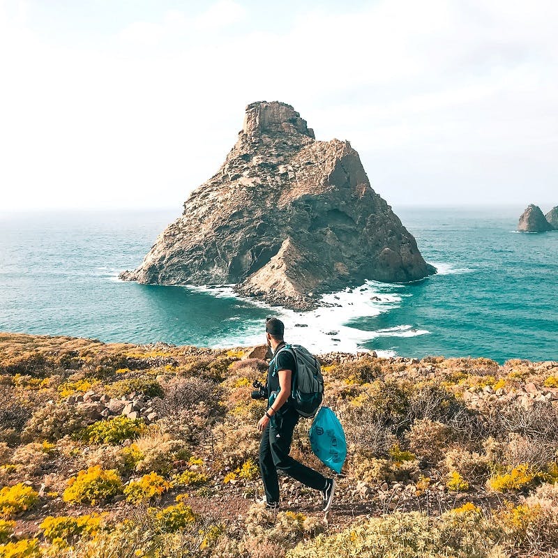 Someone carrying a rucksack and blue bag walks on the seaside with a sea-stack in the background.