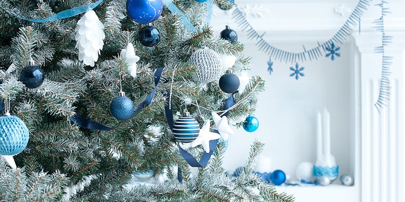 Unique holiday decor in blue and white on an evergreen tree with a snowflake garland in the background